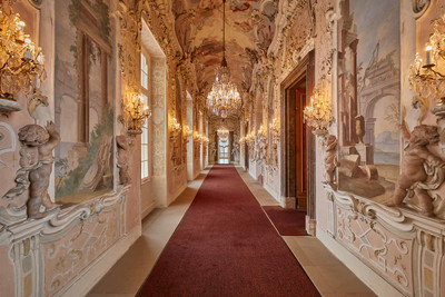 Langer Gang, ausgelegt mit einem roten Teppich im Inneren eines Schlosses. An den Wänden hängen Kronleuchter und schmucke barocke Verzierungen.